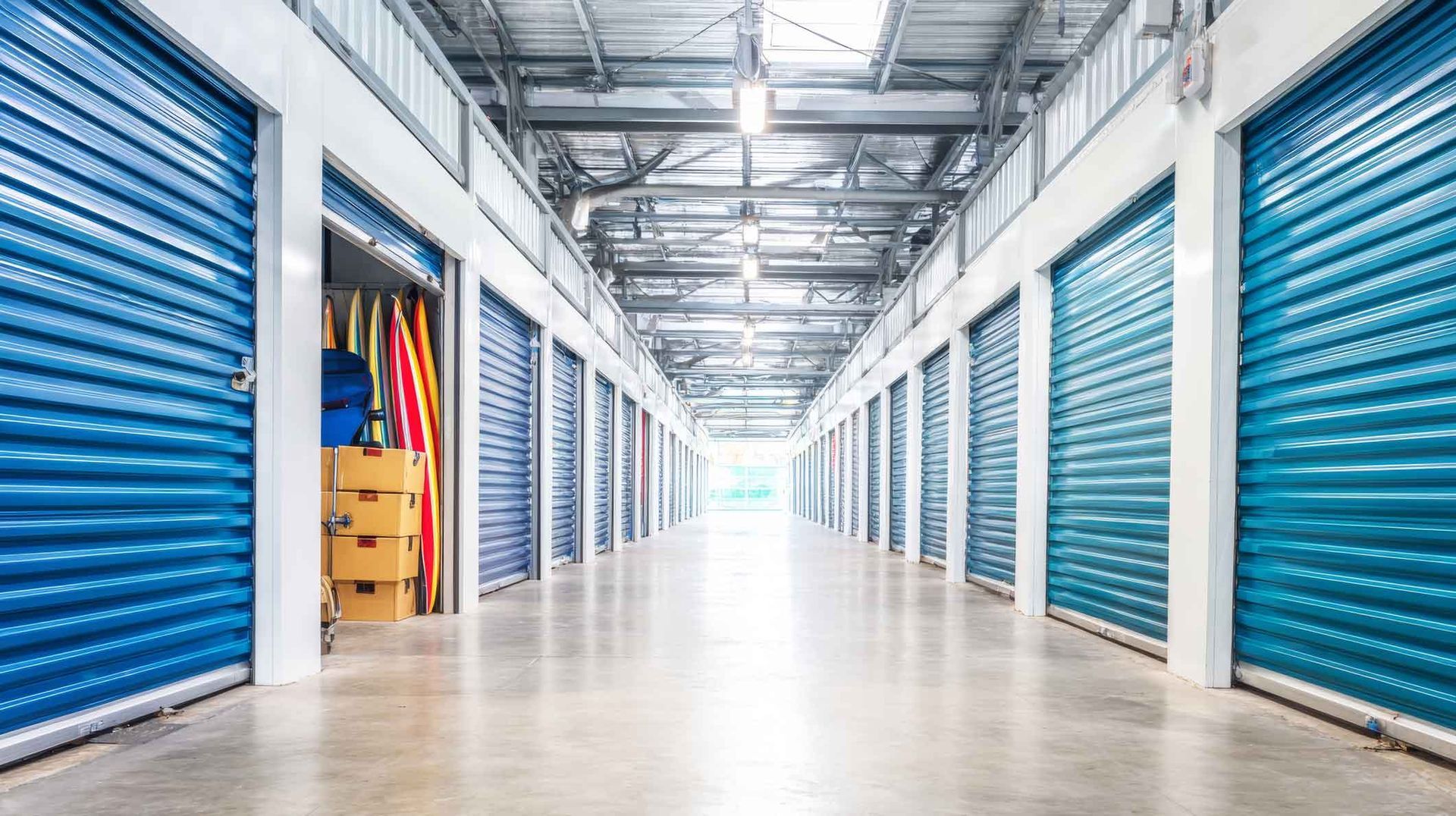 Bright hallway lined with blue roll-up doors; one open unit reveals boxes and colorful surfboards.