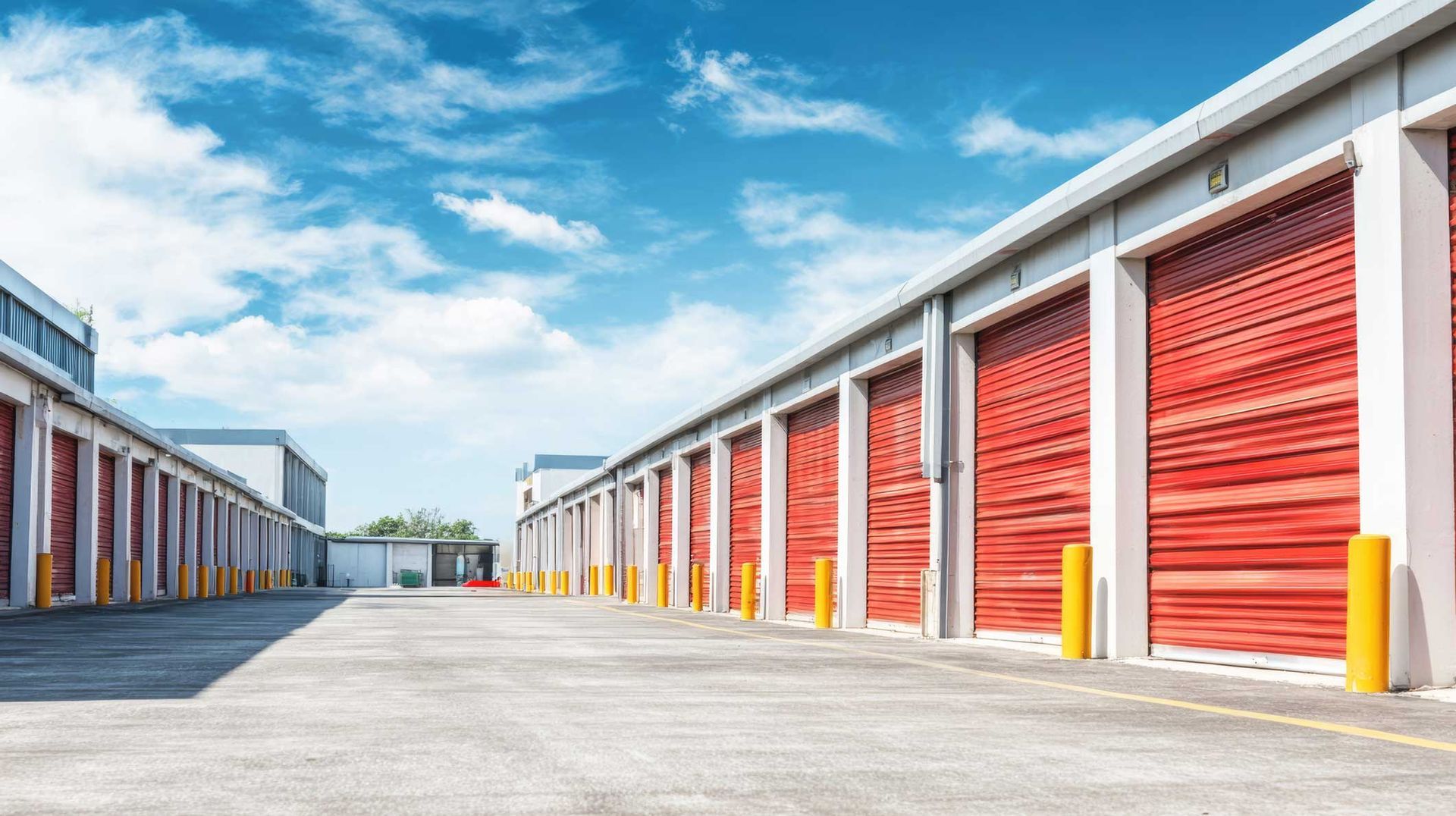 Rows of clean self-storage units with red roll-up doors, wide paved drive under blue sky outdoors.