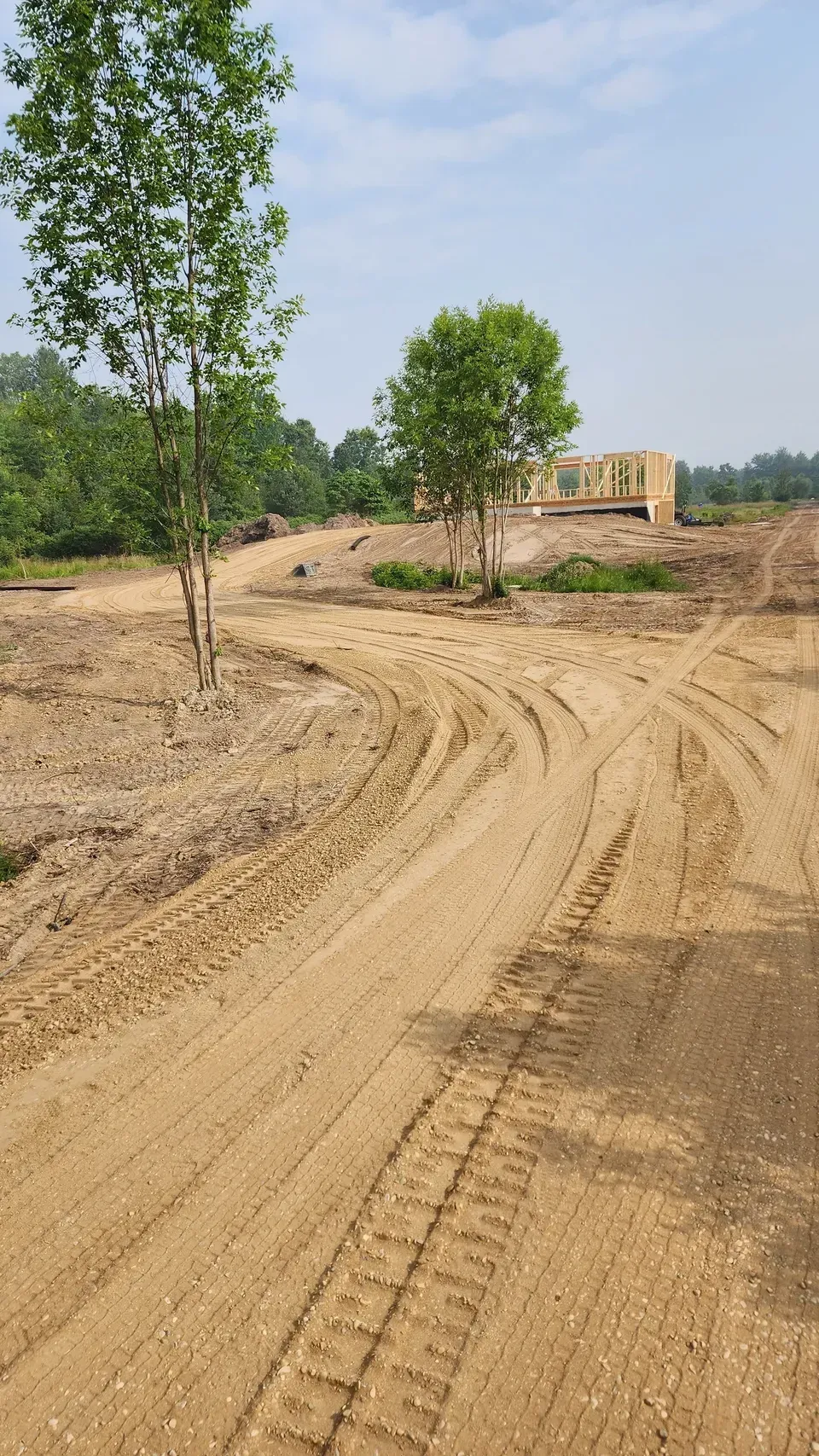 Dirt road with tire tracks, trees, and a wooden building under a blue sky.