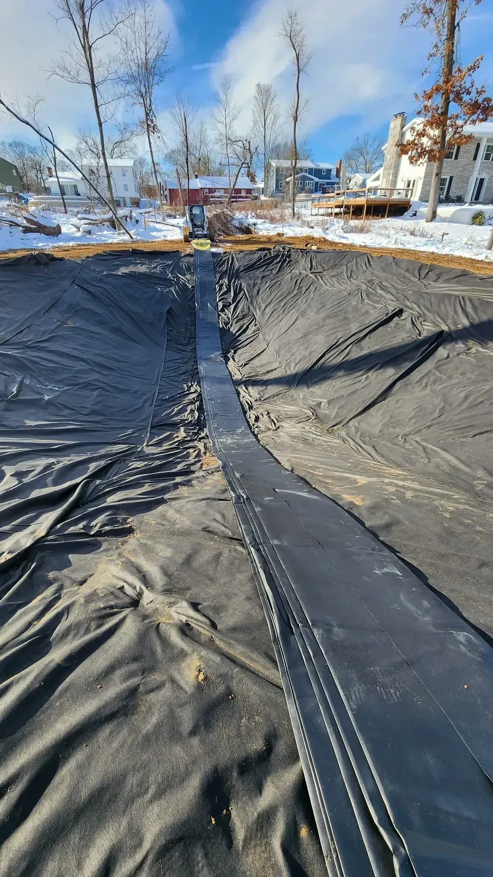 Black fabric lining a pool excavation site, with a small excavator in the background and a snow-covered landscape.