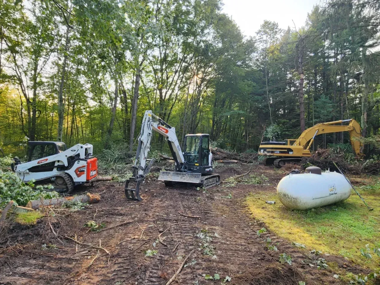 Construction site with three excavators on muddy ground. One is a Bobcat, one has a grapple arm, and another is yellow.