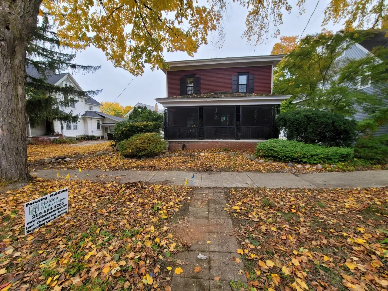 Two-story red house with a black porch, surrounded by fall foliage.