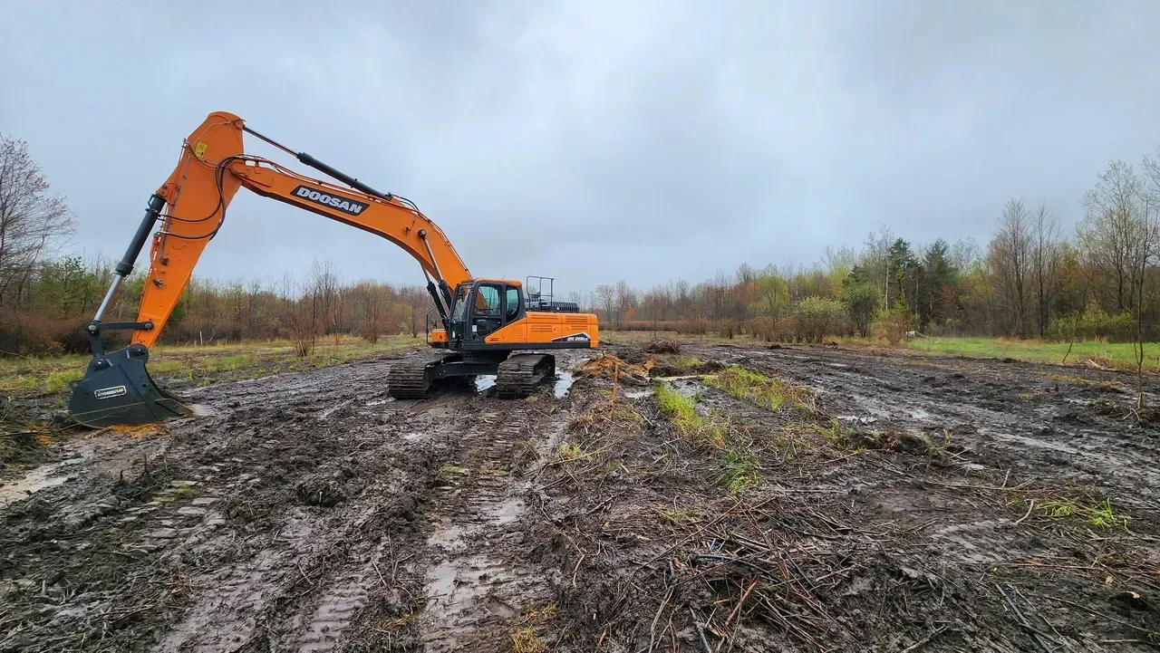 Orange excavator clearing a muddy field of vegetation under an overcast sky.