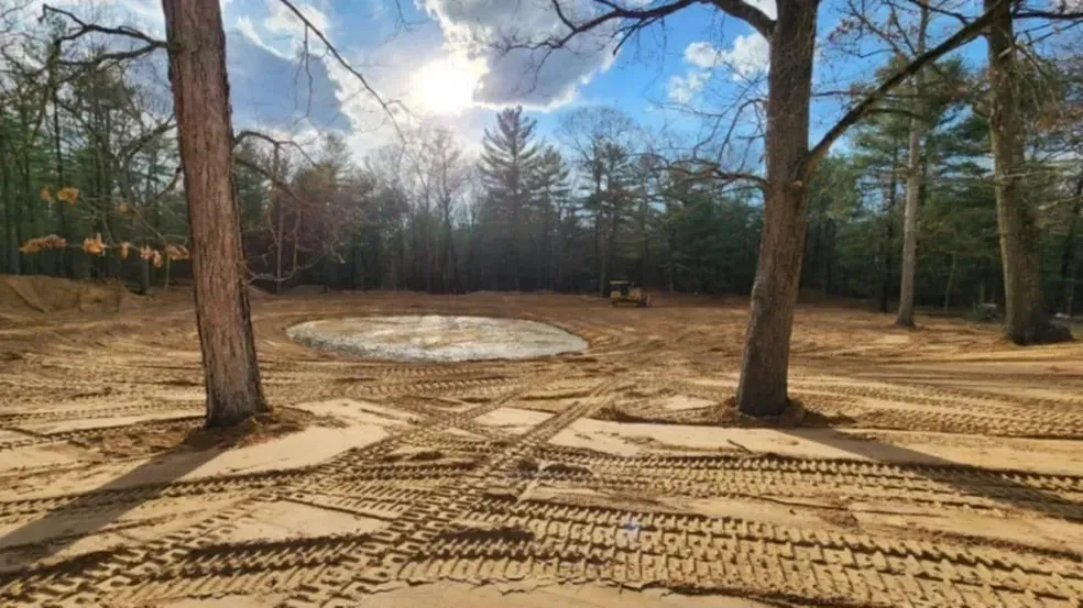 Open area with tire tracks, trees, and muddy patch under a cloudy blue sky.