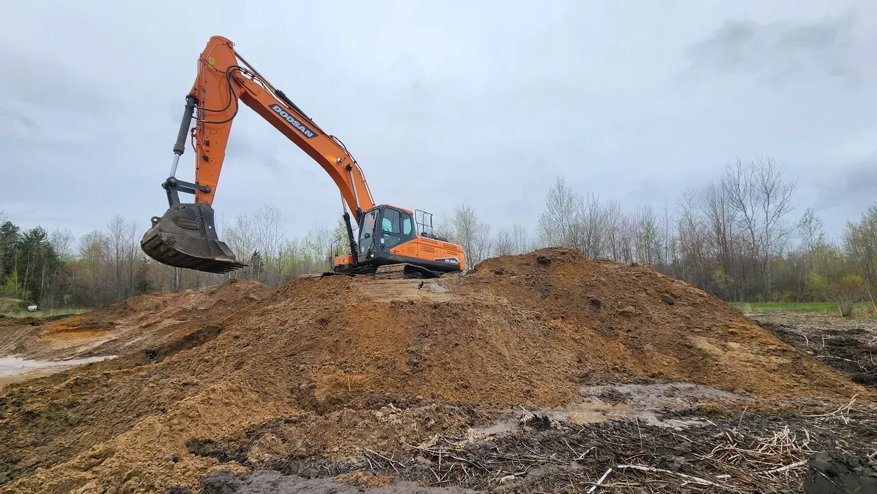 An orange excavator atop a brown dirt pile, under a cloudy sky.