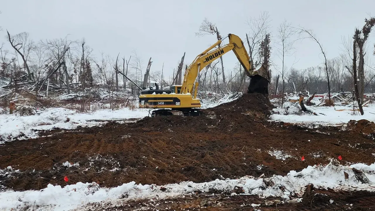 Yellow excavator digging in snowy, muddy field, trees in the background.