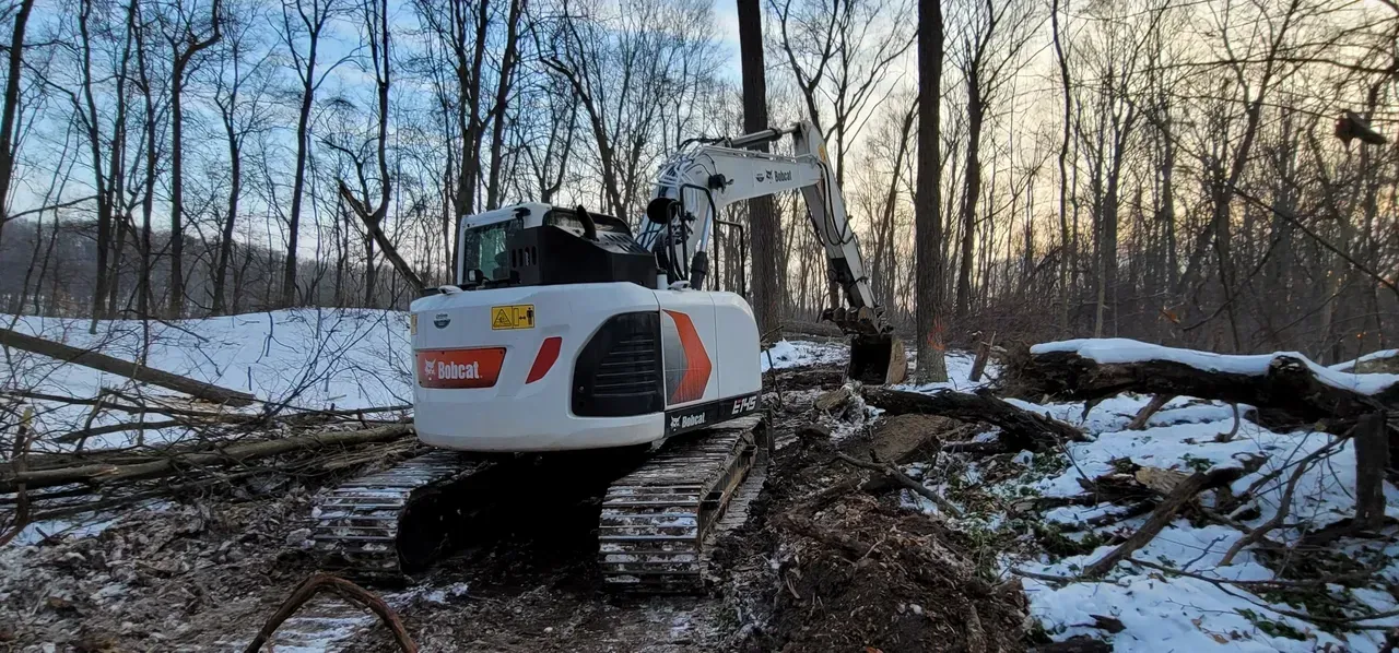 A white Bobcat excavator clearing fallen trees in a snowy forest.