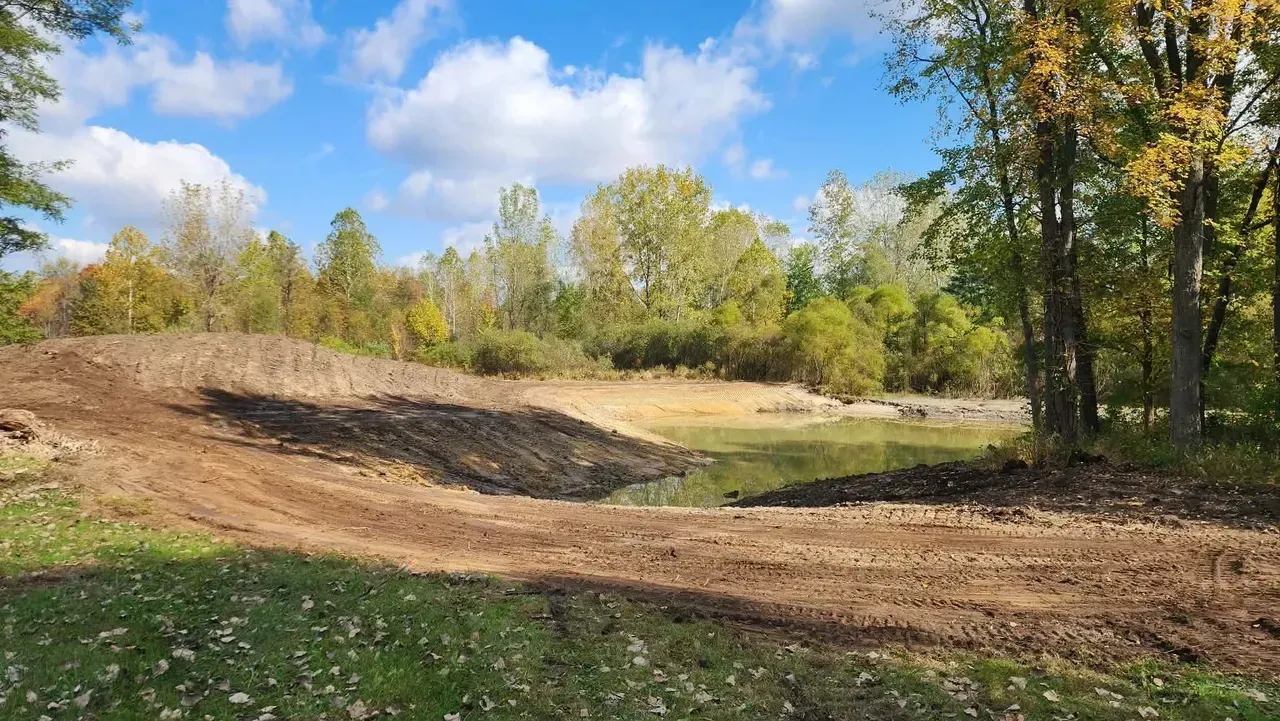 Dirt embankment next to a small pond, surrounded by trees with autumn foliage, under a blue sky.