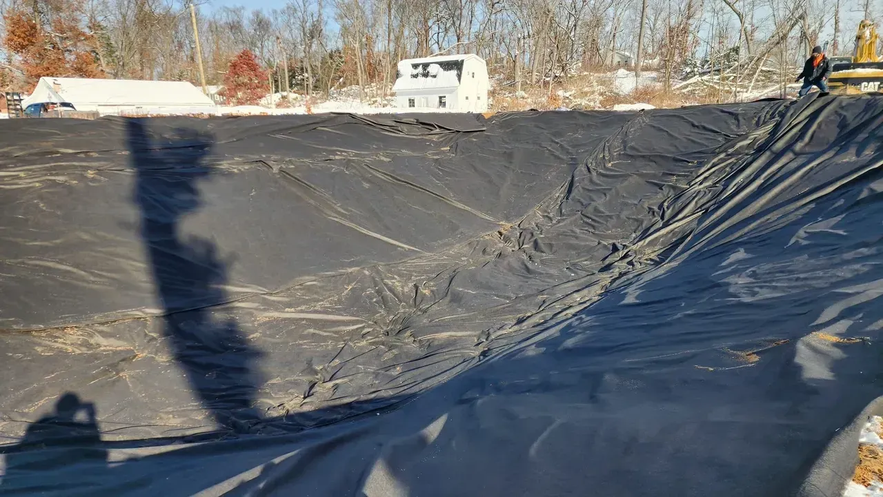 Black liner covering a large, excavated area. A person and machinery on the edge, snow in the background.
