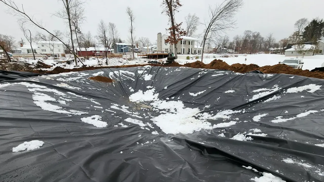 Black liner in a dug-out area, partially covered in snow. Houses and trees in the background.