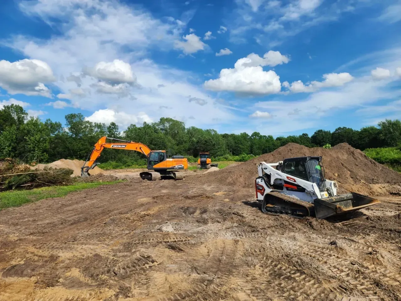 Construction site with excavator and skid steer on a dirt field under a cloudy blue sky.