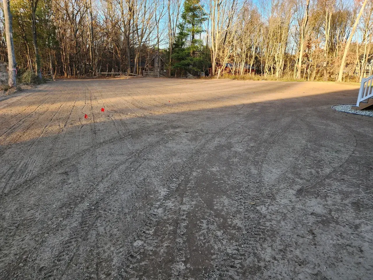 Gravel-covered lot with stakes, sunlight, and a backdrop of trees.