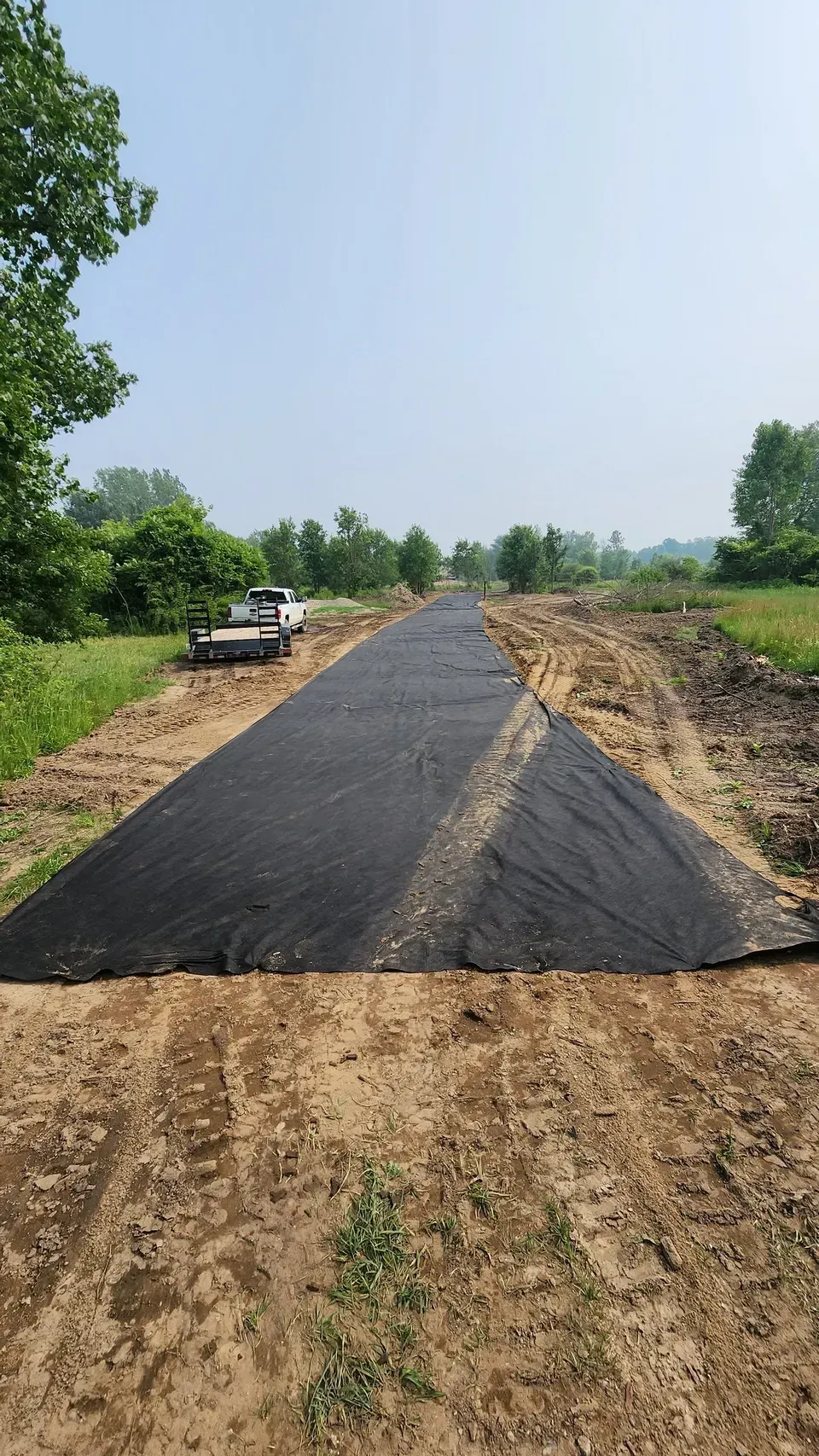 Dirt road with a large black fabric covering a portion of it, trees in the background, blue sky.