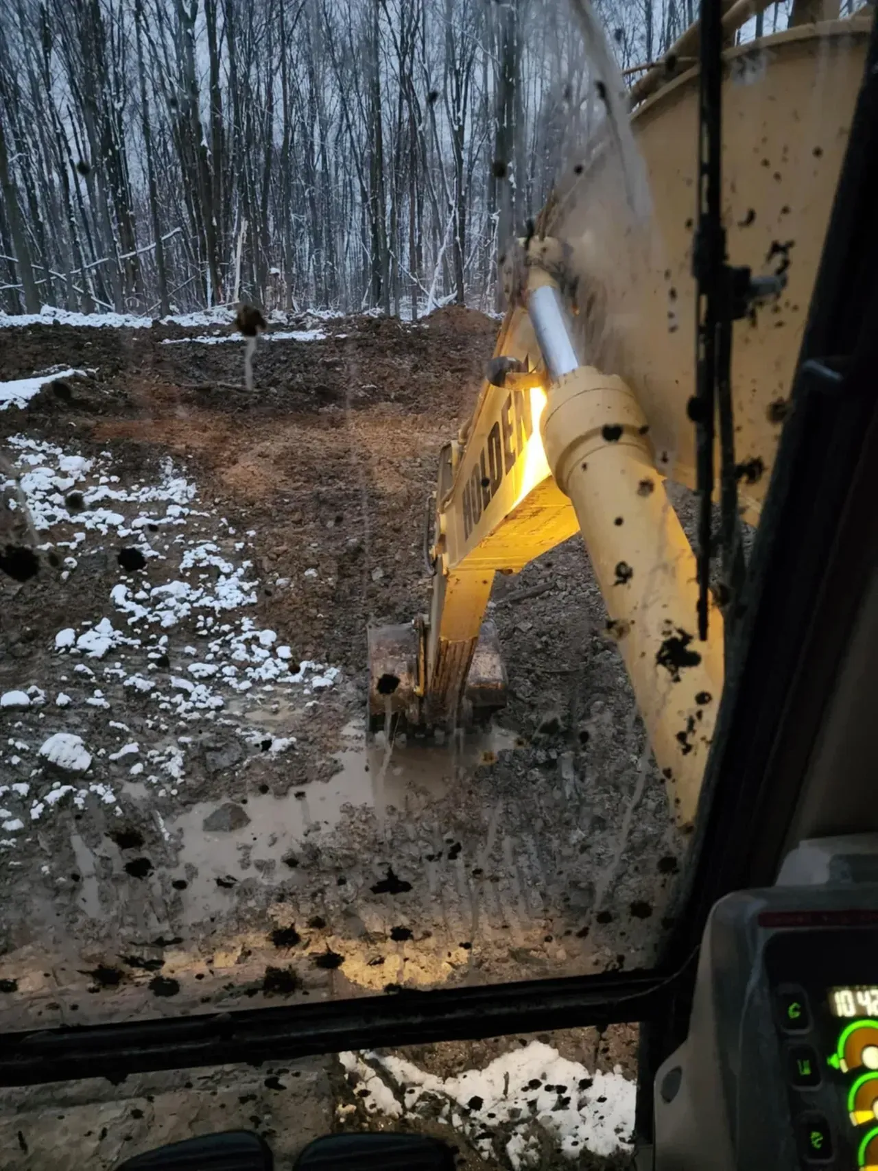Muddy excavator arm scooping dirt; seen through a dirty window, forest in background.