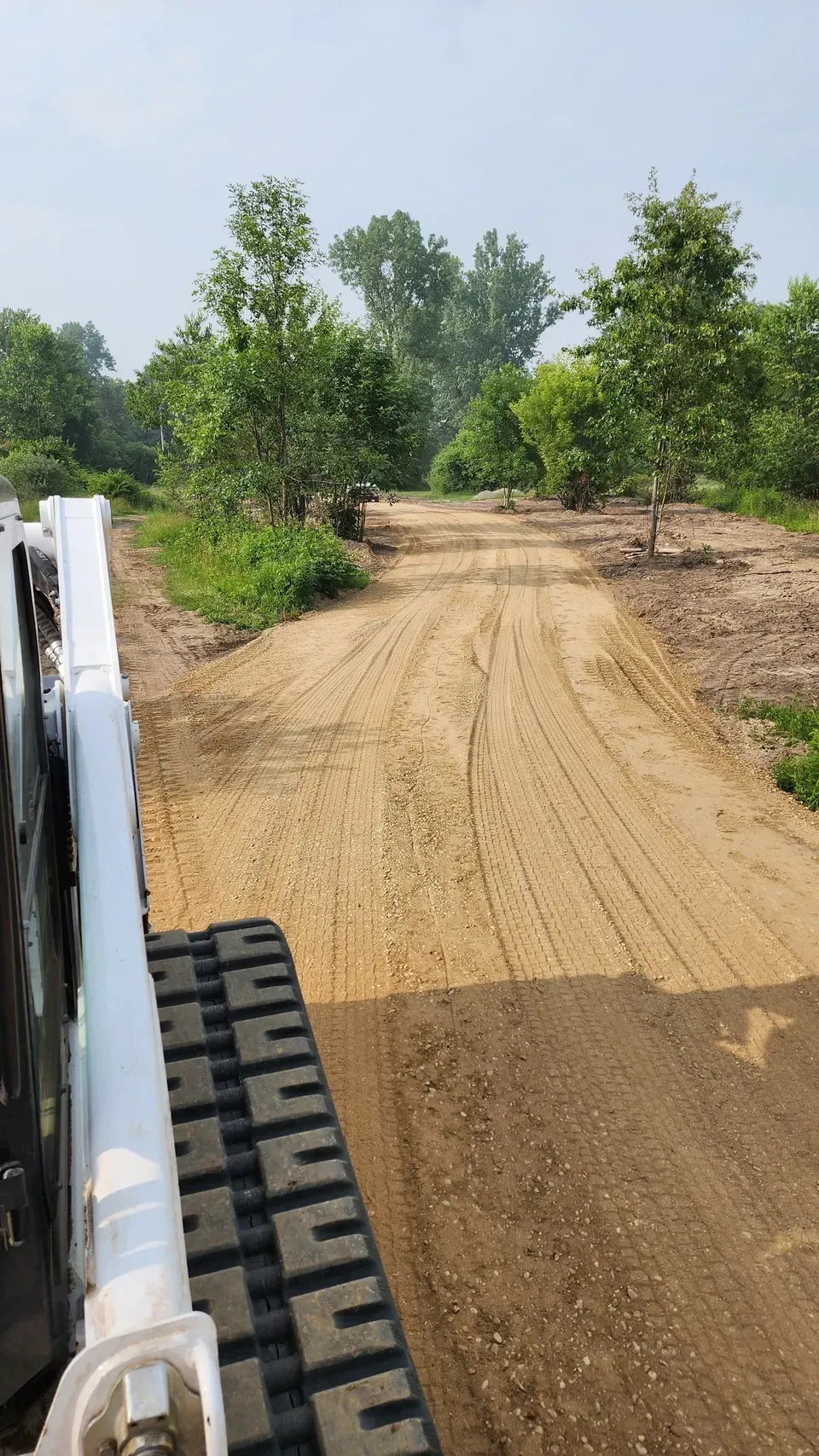 Bobcat leveling a gravel road, trees in the background under a blue sky.