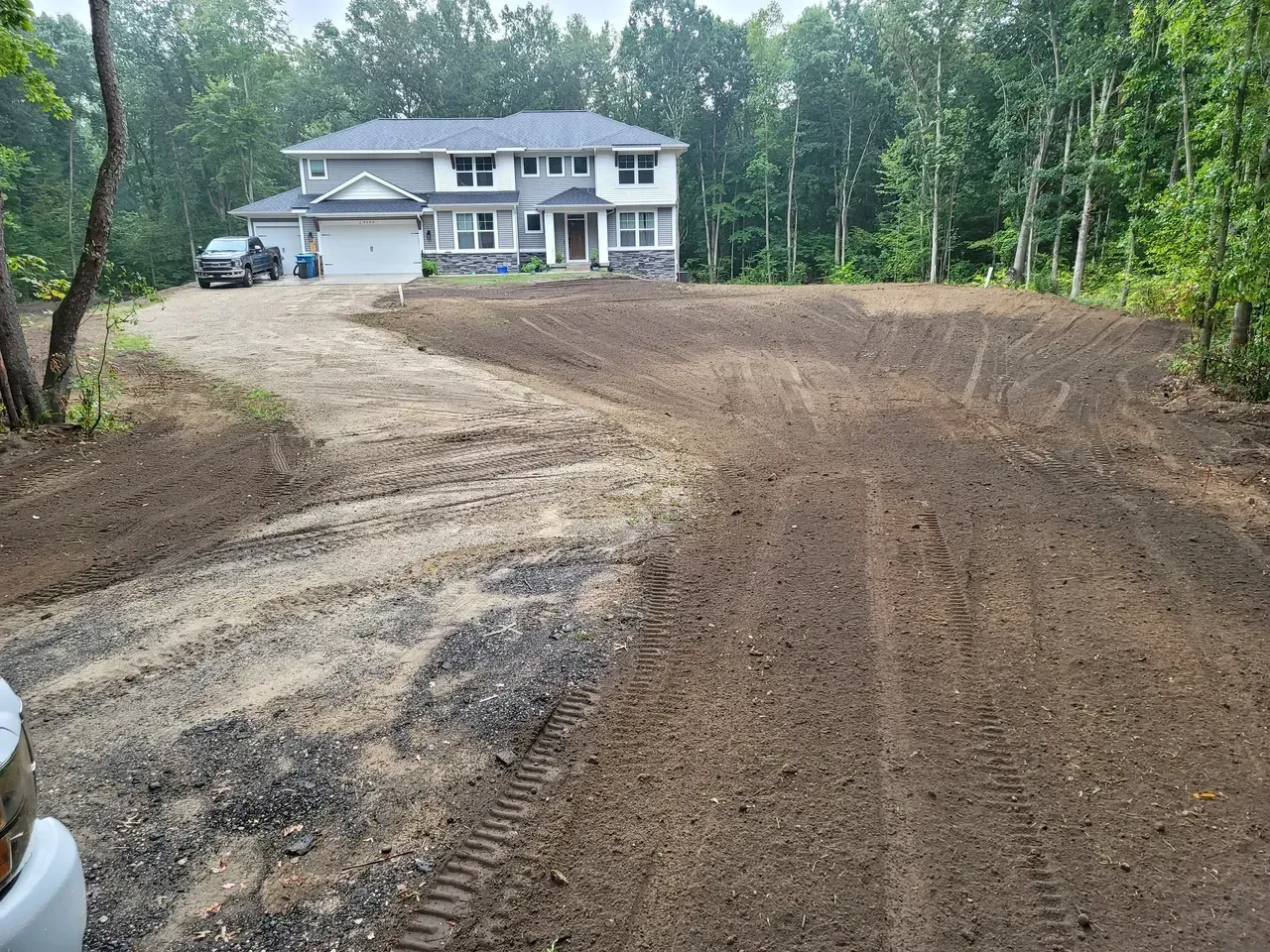 Driveway leading to a two-story house surrounded by trees. Landscaping work in progress.