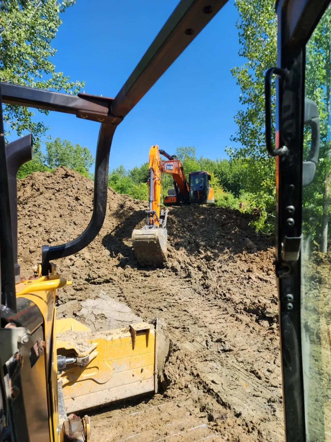 View from a bulldozer cab; two excavators work on a muddy slope under a blue sky, surrounded by trees.