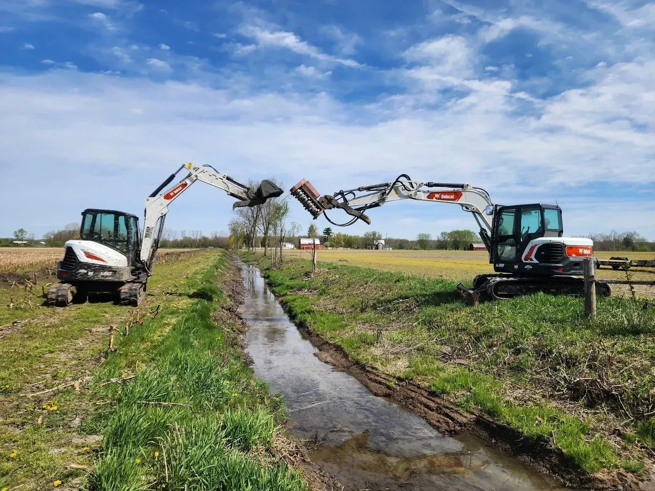 Two Bobcat excavators working in a drainage ditch in a field. Blue sky.
