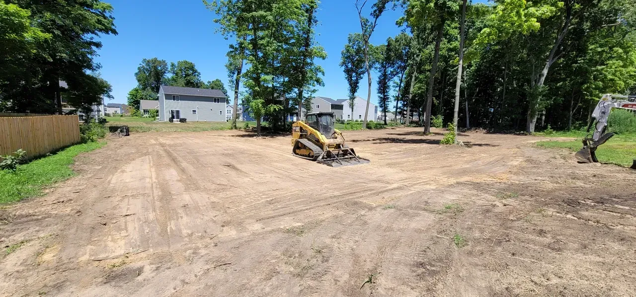 A skid steer tractor clearing a wooded lot, trees in the background, blue sky.