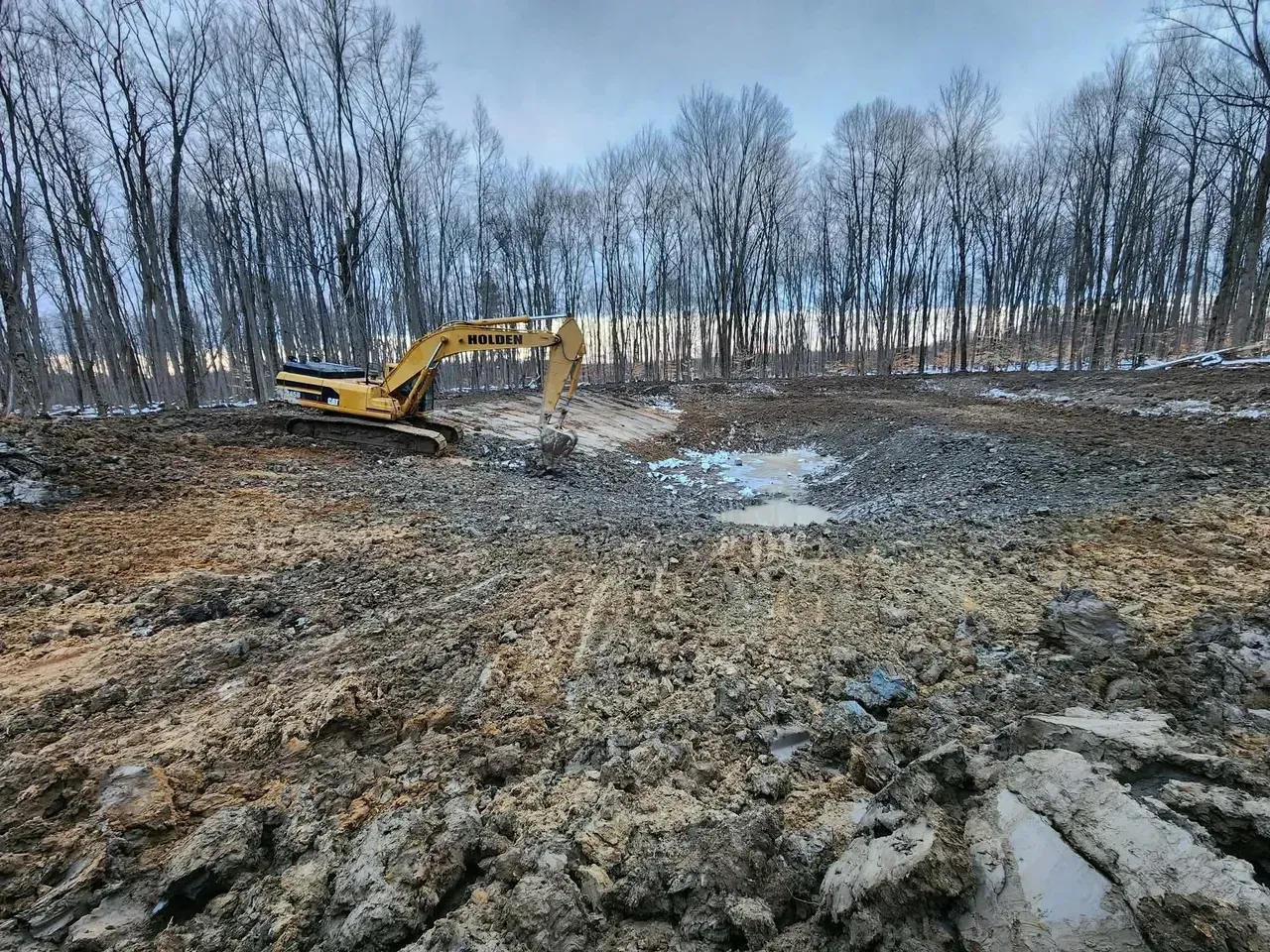 Yellow excavator digs in muddy ground, surrounded by trees with frost. Overcast sky.