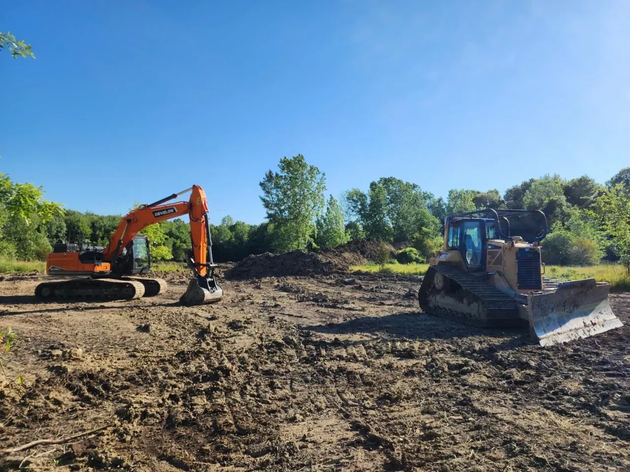 An excavator and bulldozer clearing land in front of a forest under a blue sky.