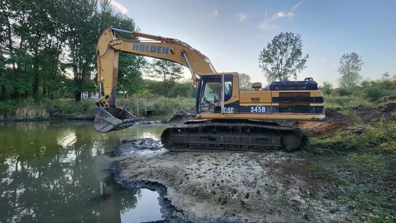 Yellow excavator working in a murky pond, digging near a treeline.