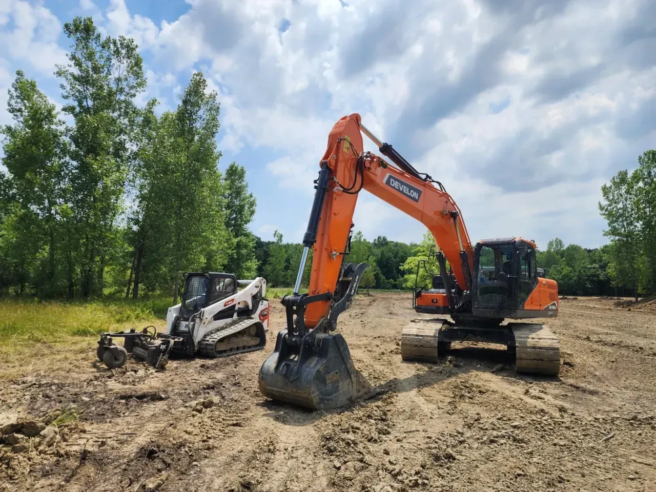 Construction site with an orange excavator and a white skid steer on a cleared area, surrounded by trees and a cloudy sky.