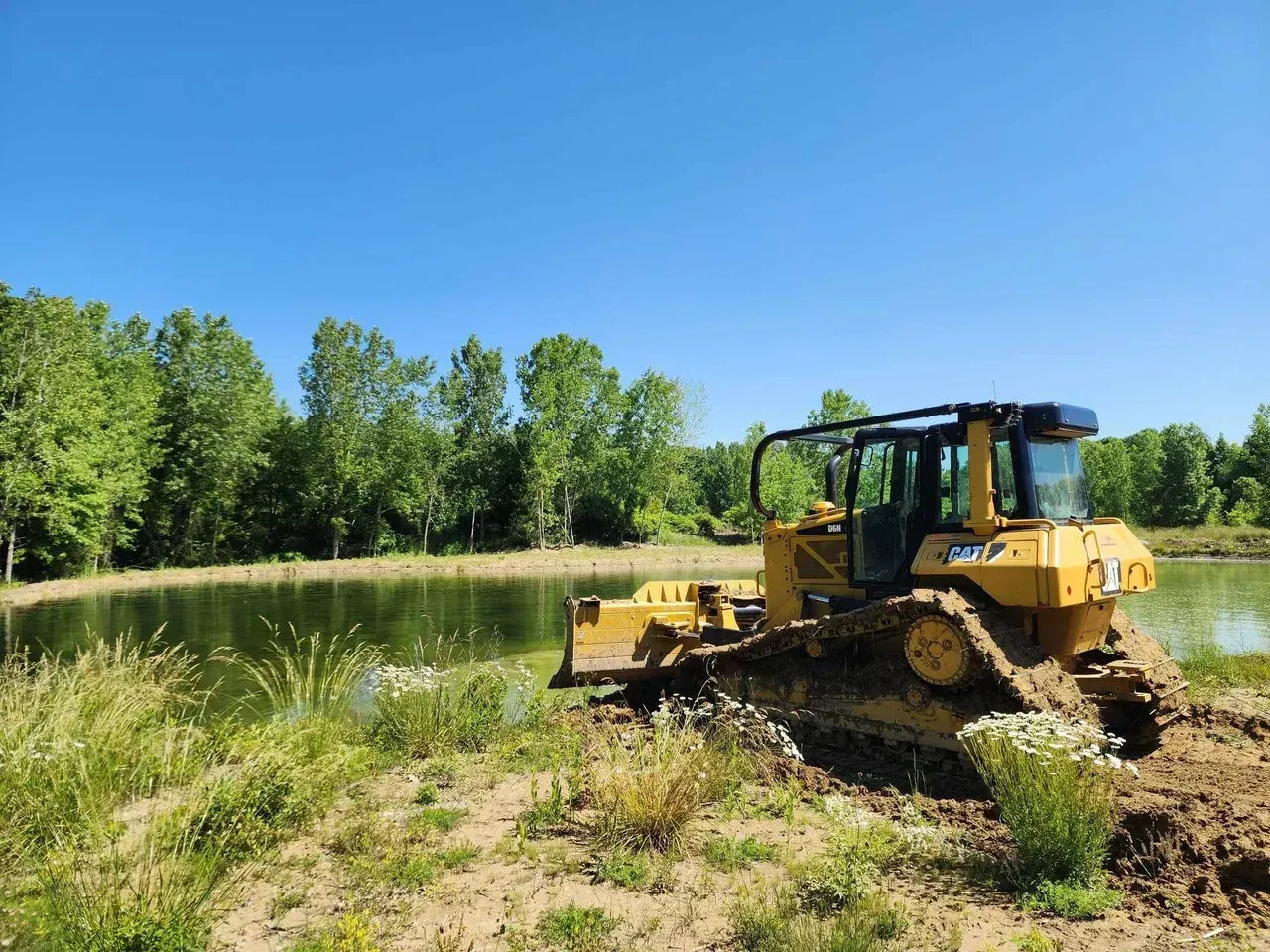 Yellow bulldozer on muddy ground next to a pond. Green trees and blue sky in the background.