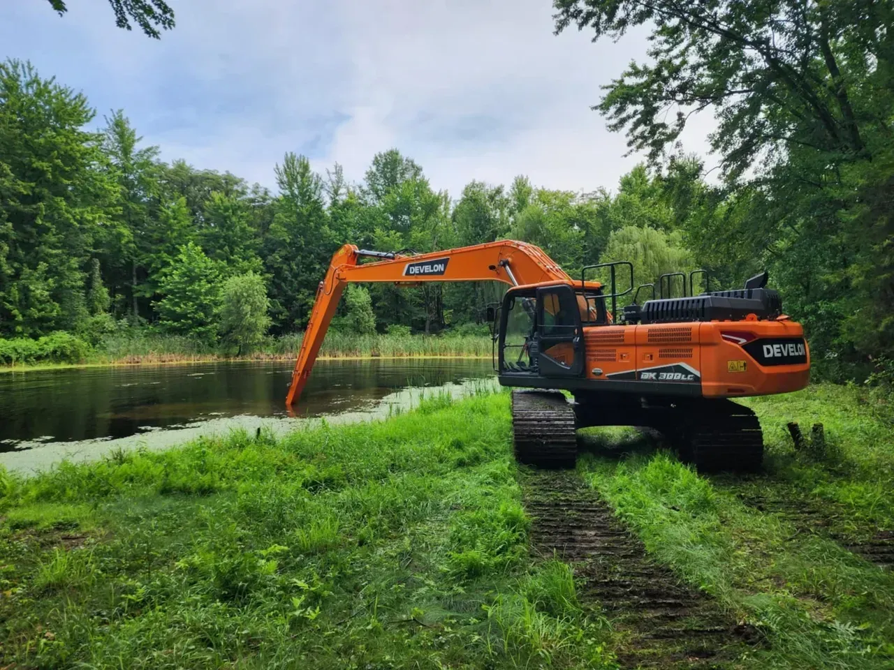 Orange excavator in a pond, digging near lush green vegetation and trees. Cloudy sky.