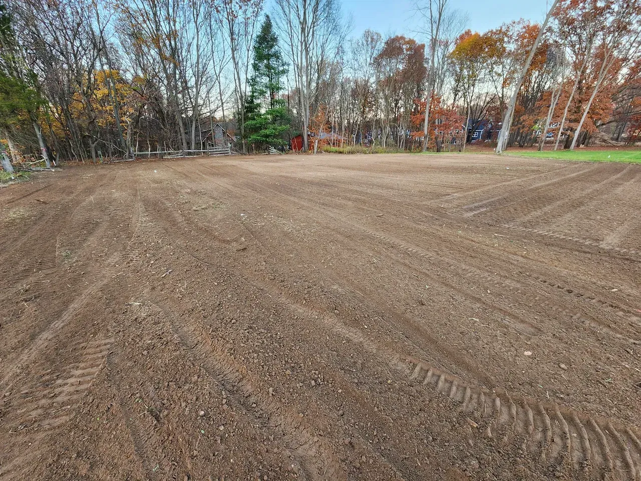 Tilled brown earth field, with tire tracks, surrounded by trees with autumn foliage.
