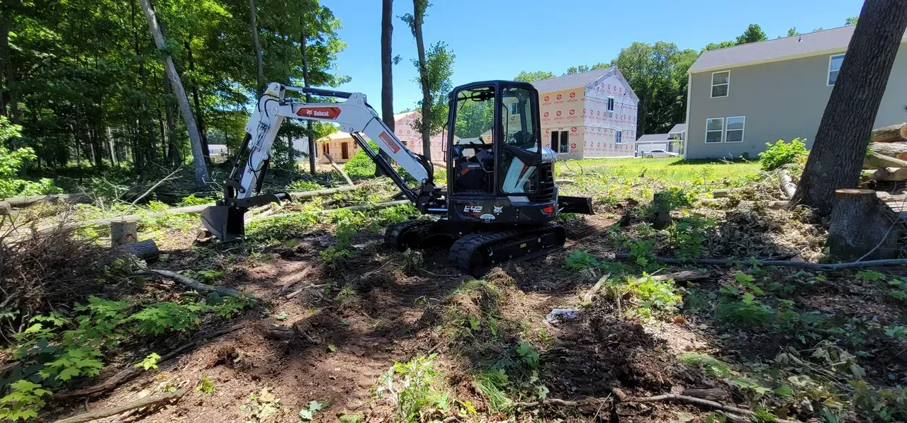 A Bobcat excavator clearing brush in a wooded area near houses on a sunny day.