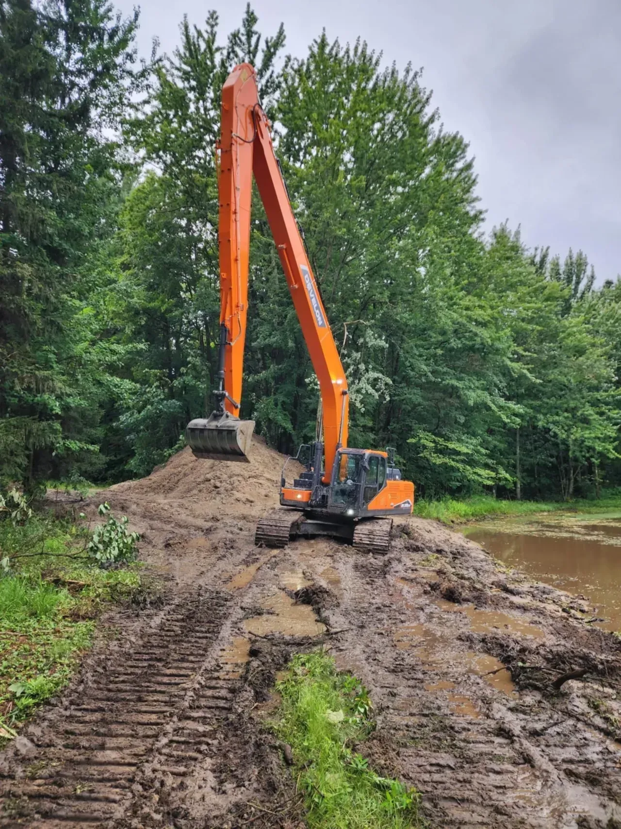 Orange excavator on muddy ground near a pond and green trees.