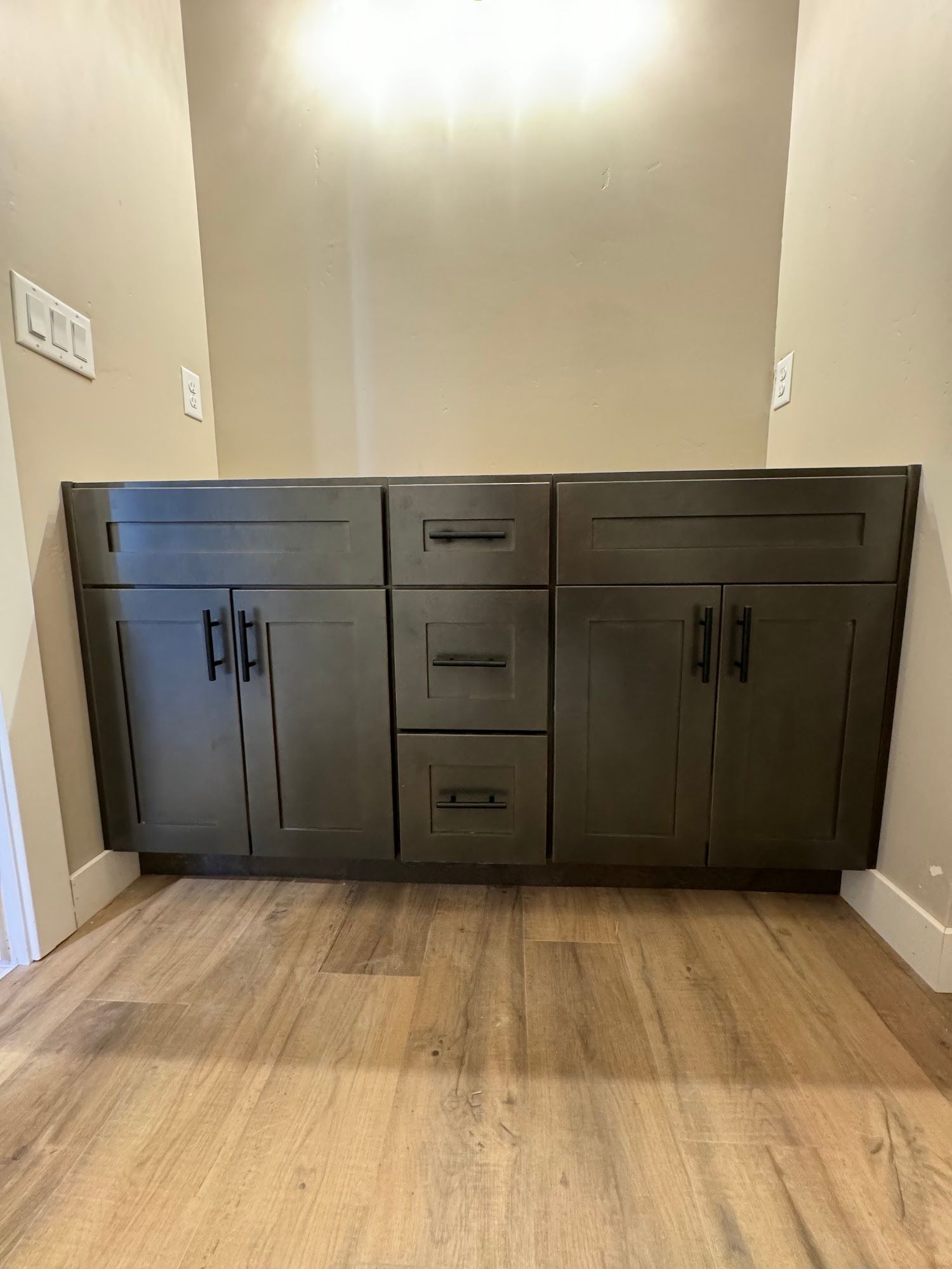 Gray bathroom vanity with cabinets and drawers, black hardware, against beige wall, on wood-look flooring.