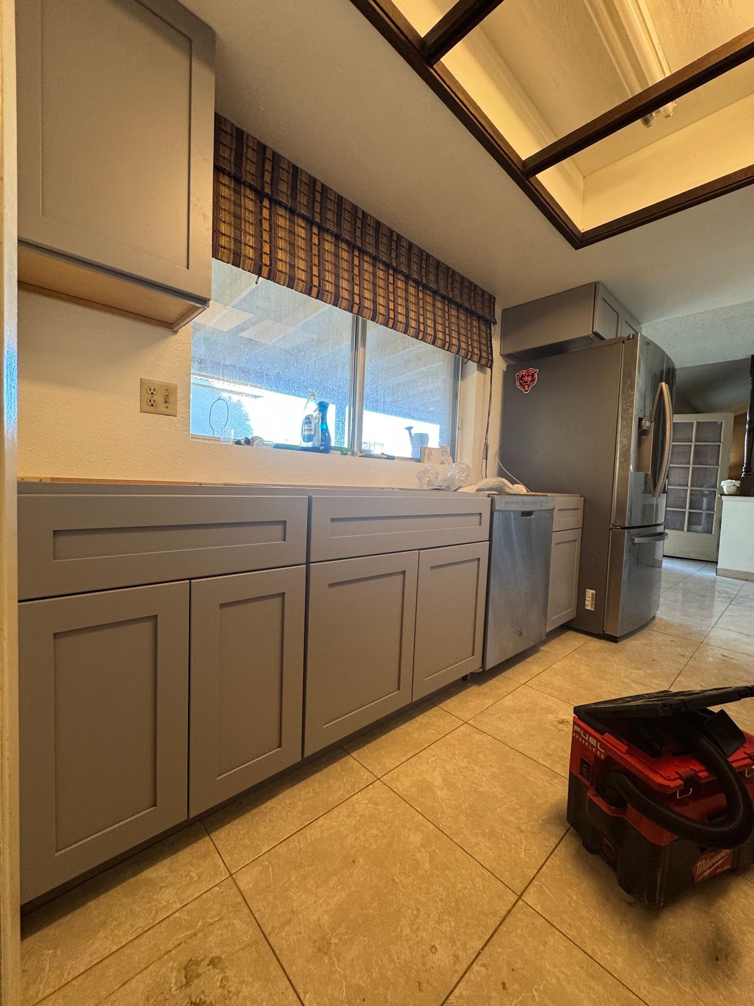 Gray kitchen cabinets under a window with a brown blind; stainless steel refrigerator.