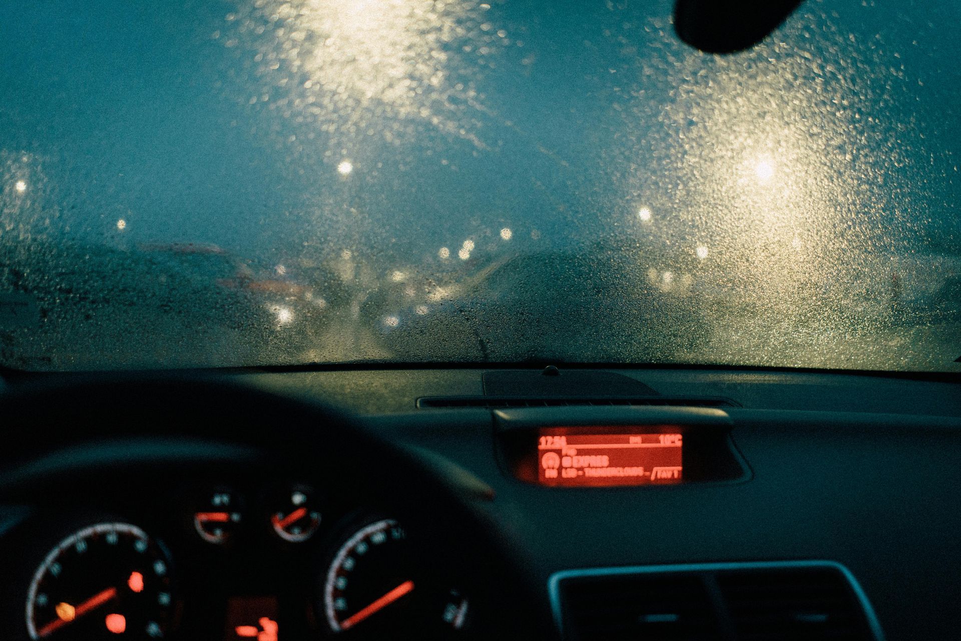 A rain-streaked car windshield at night, with blurred streetlights visible beyond the dashboard.