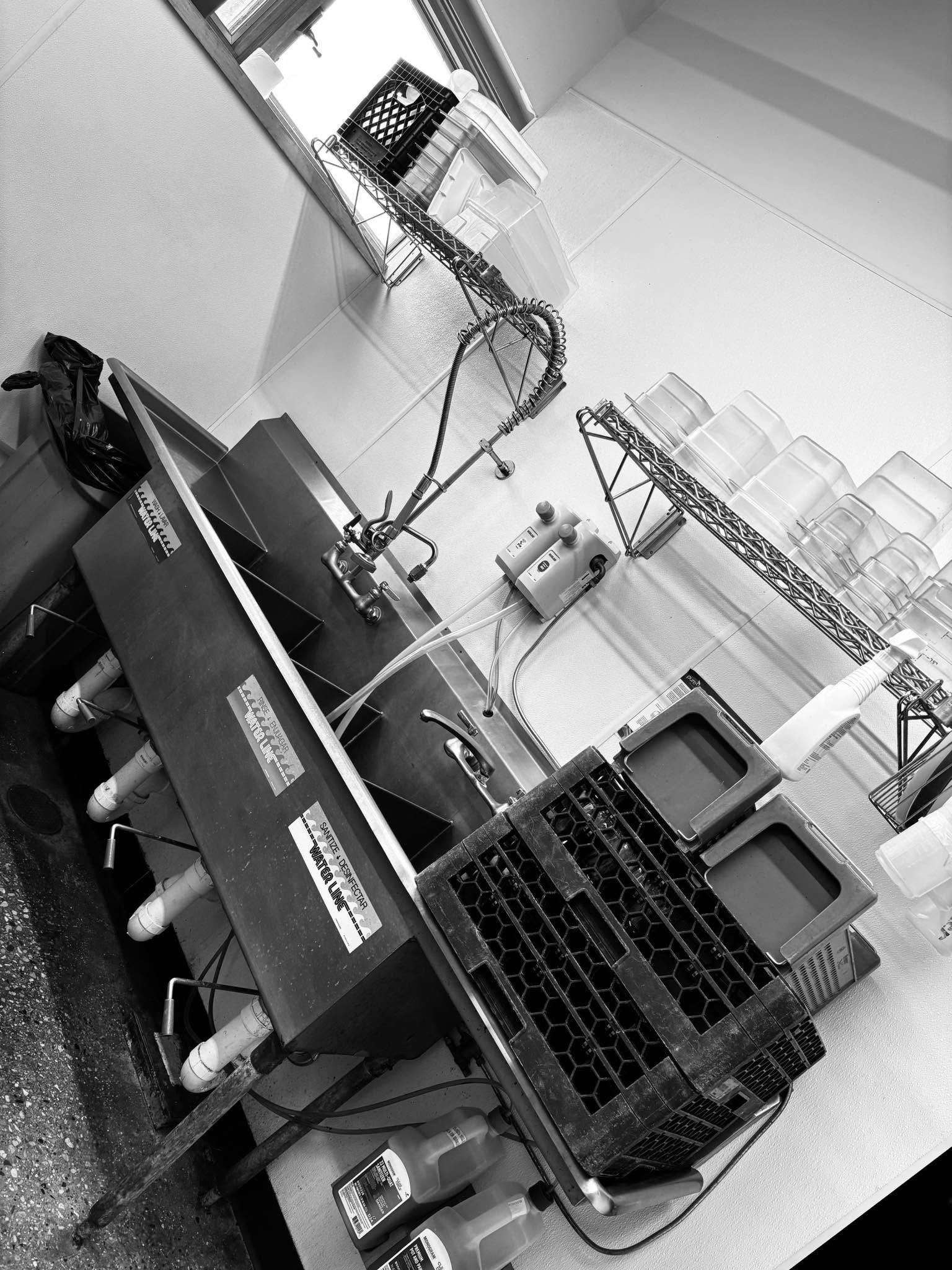 Tilted black-and-white photo of a workshop with shelves, equipment, and a monitor on a desk.