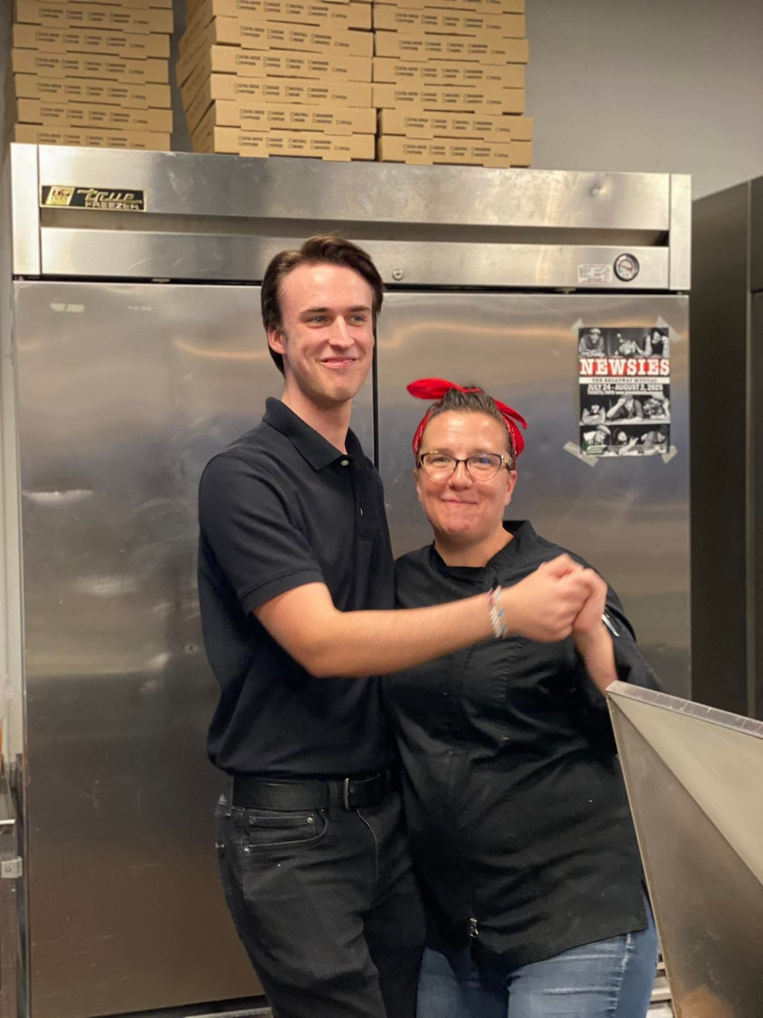 Two people in black shirts smiling and embracing in a stainless-steel kitchen area.