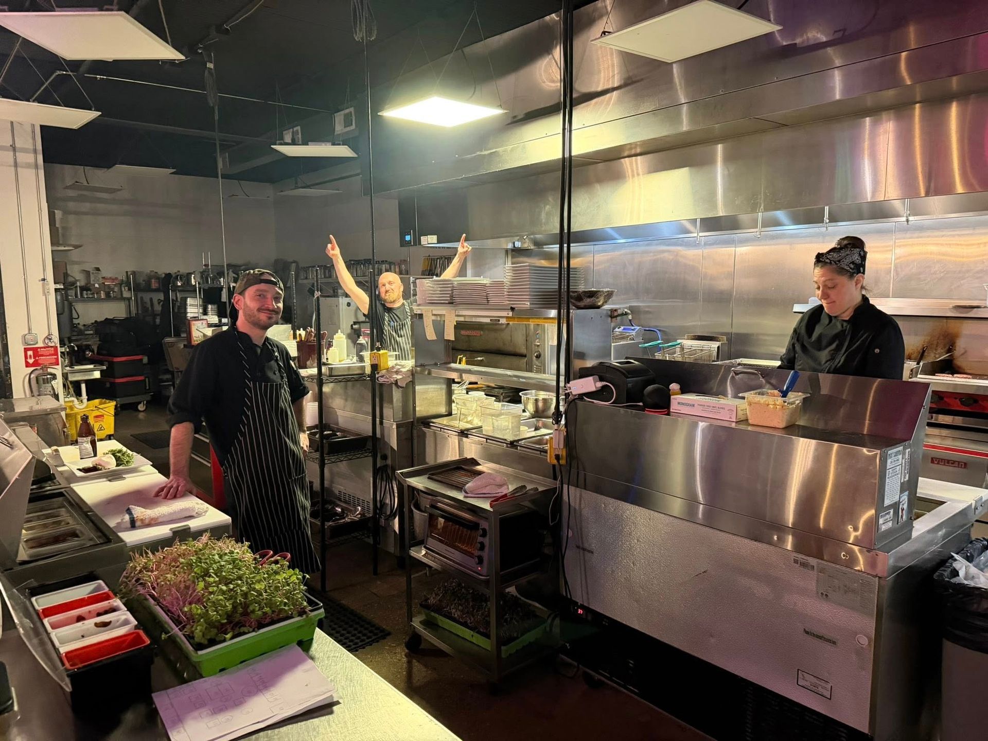 Three restaurant staff in a commercial kitchen; one raises arms, others work. Stainless steel counters, bright lights.