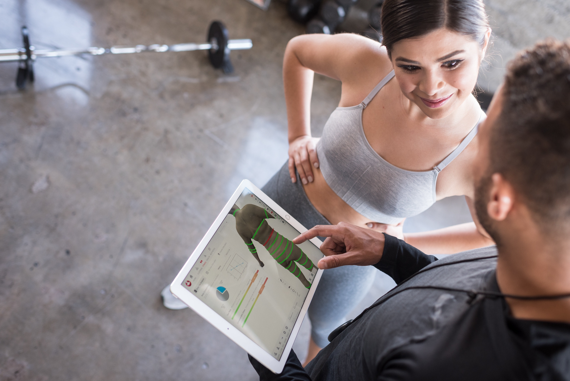 A woman in workout clothes looking at a tablet held by a man in a gym, pointing.