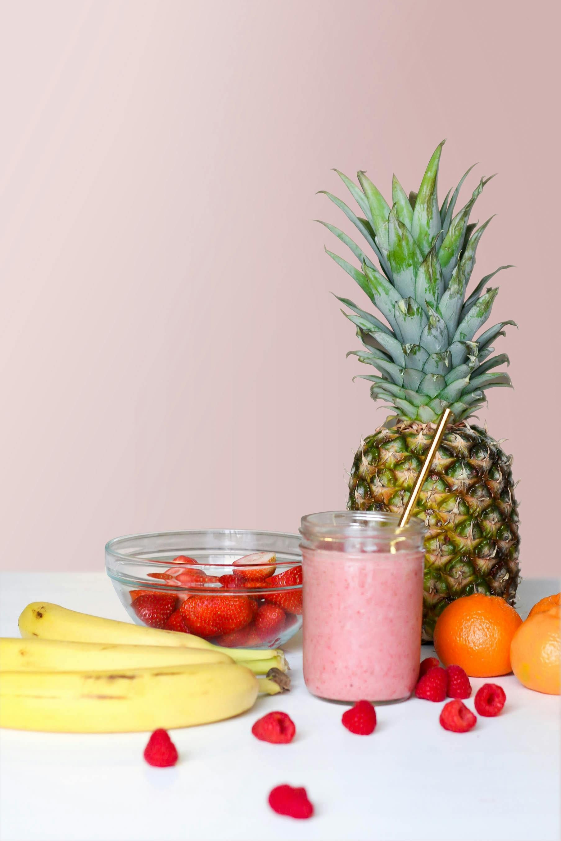 Pink smoothie in a jar with various fruits including banana, pineapple, strawberries, and oranges on a white surface.