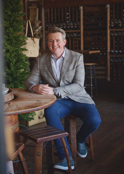 Man seated at a wooden table in a wine cellar, smiling in a light gray blazer and blue pants.