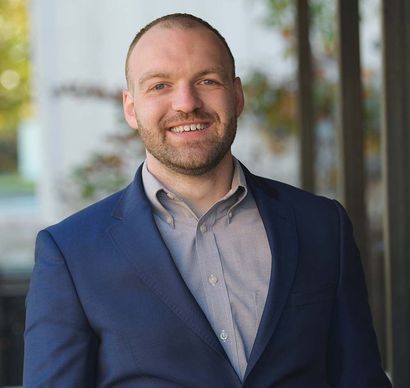 Smiling man in a navy blazer and gray shirt standing outdoors by a building