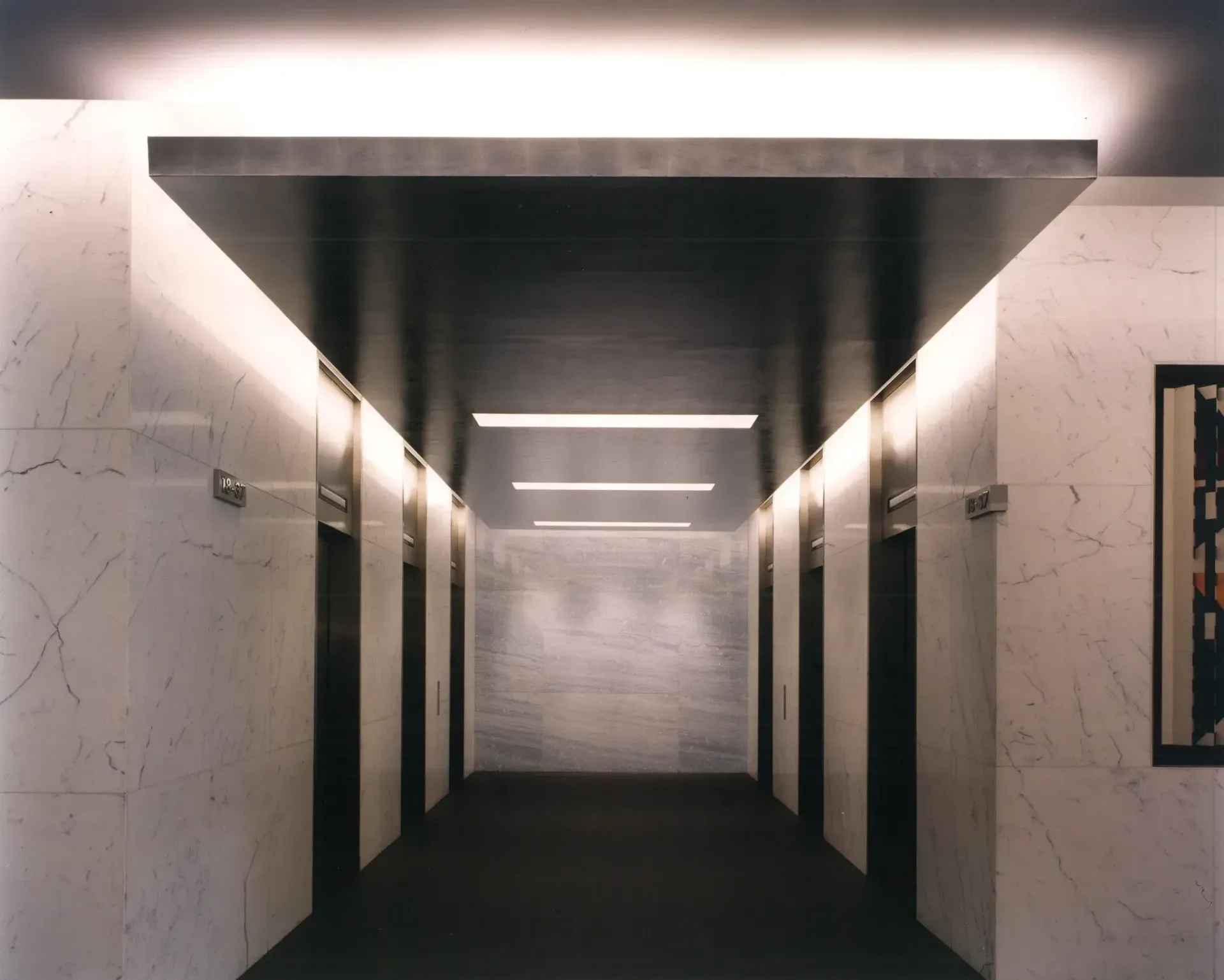A long elevator hallway with marble walls, black elevator doors, and a dark ceiling with recessed lighting.