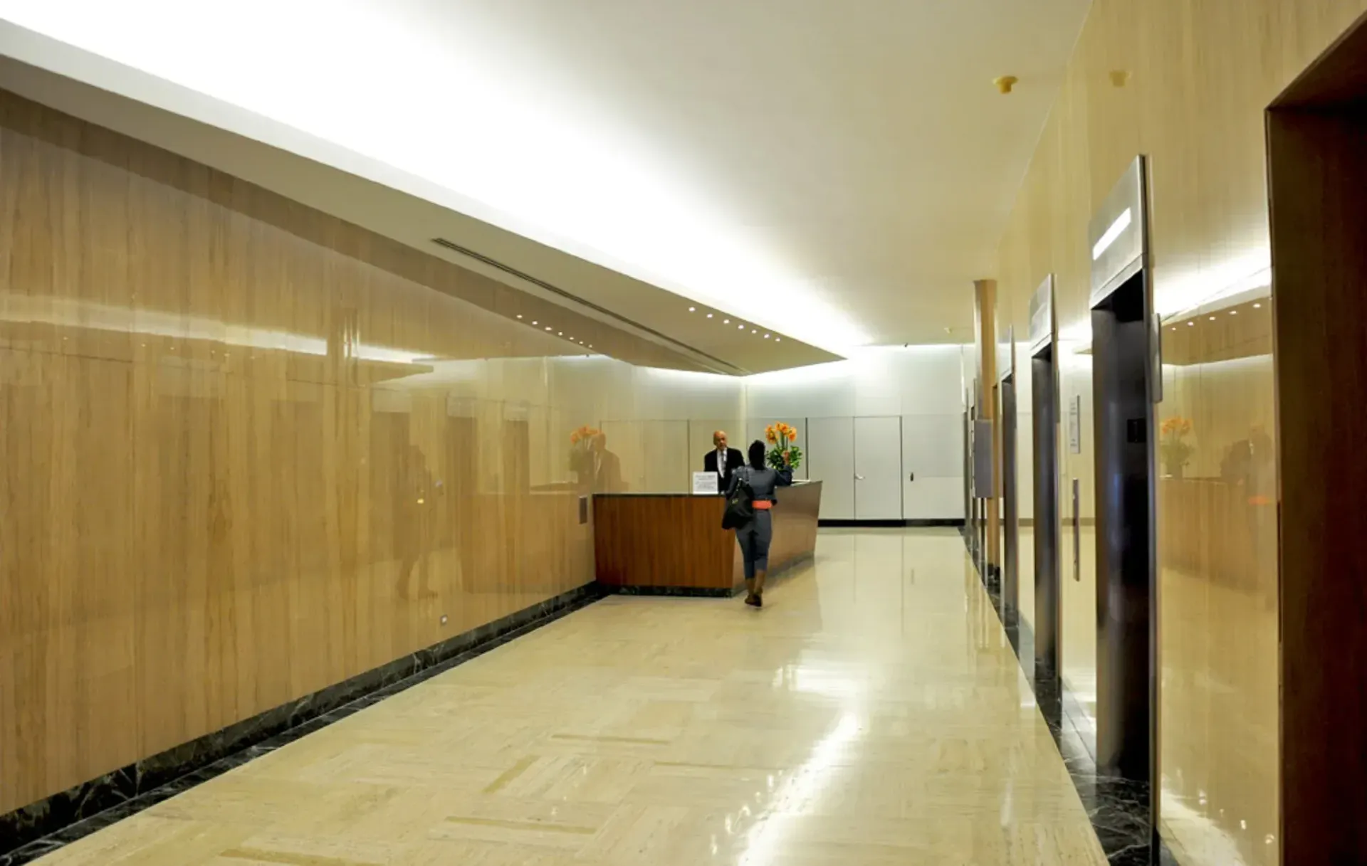 A hallway in a building with wood-paneled walls, marble floors, and elevators. A person walks toward the reception desk.