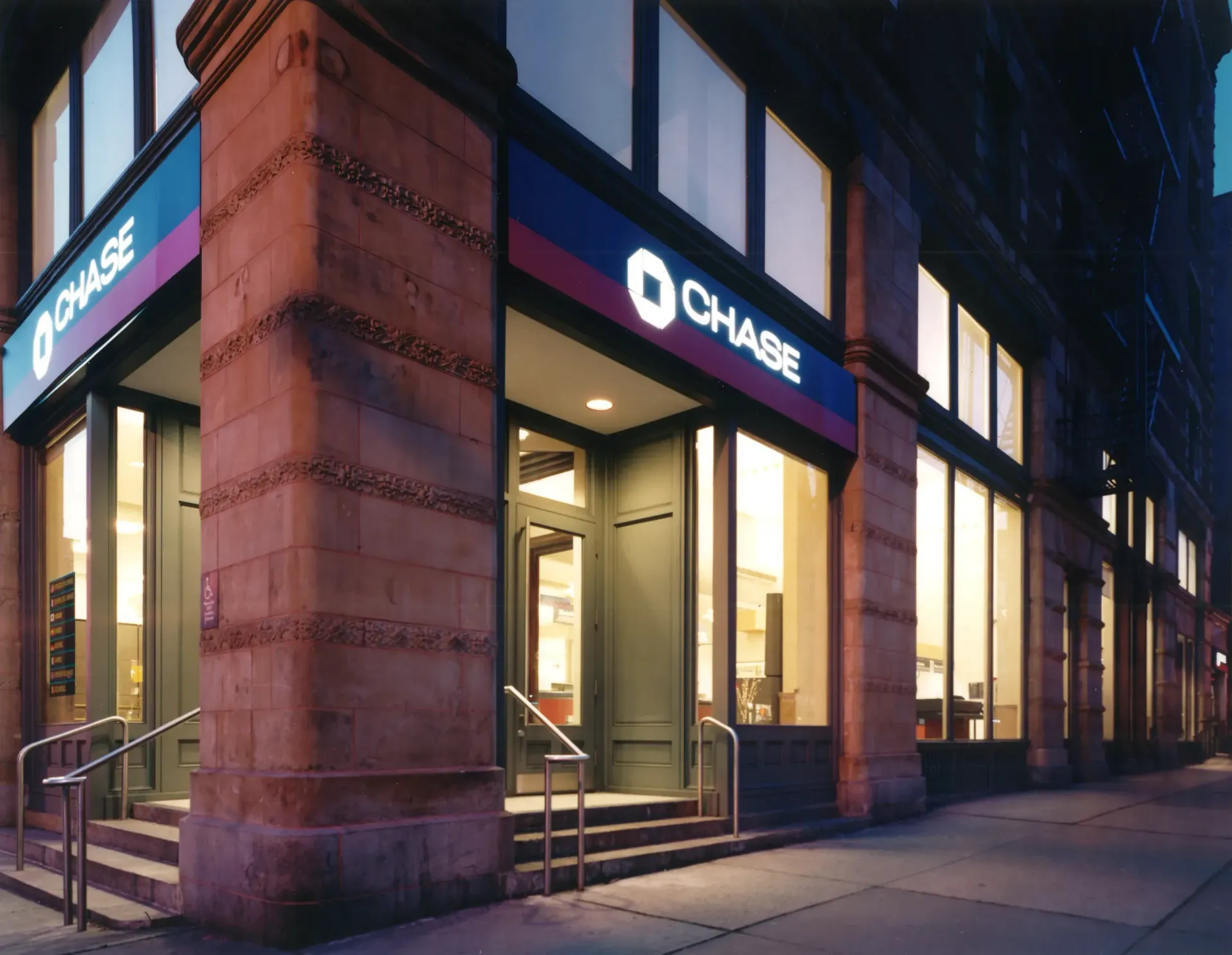 Chase bank branch on a city corner, with a red brick facade, blue and red Chase logo over entrance, and illuminated windows.