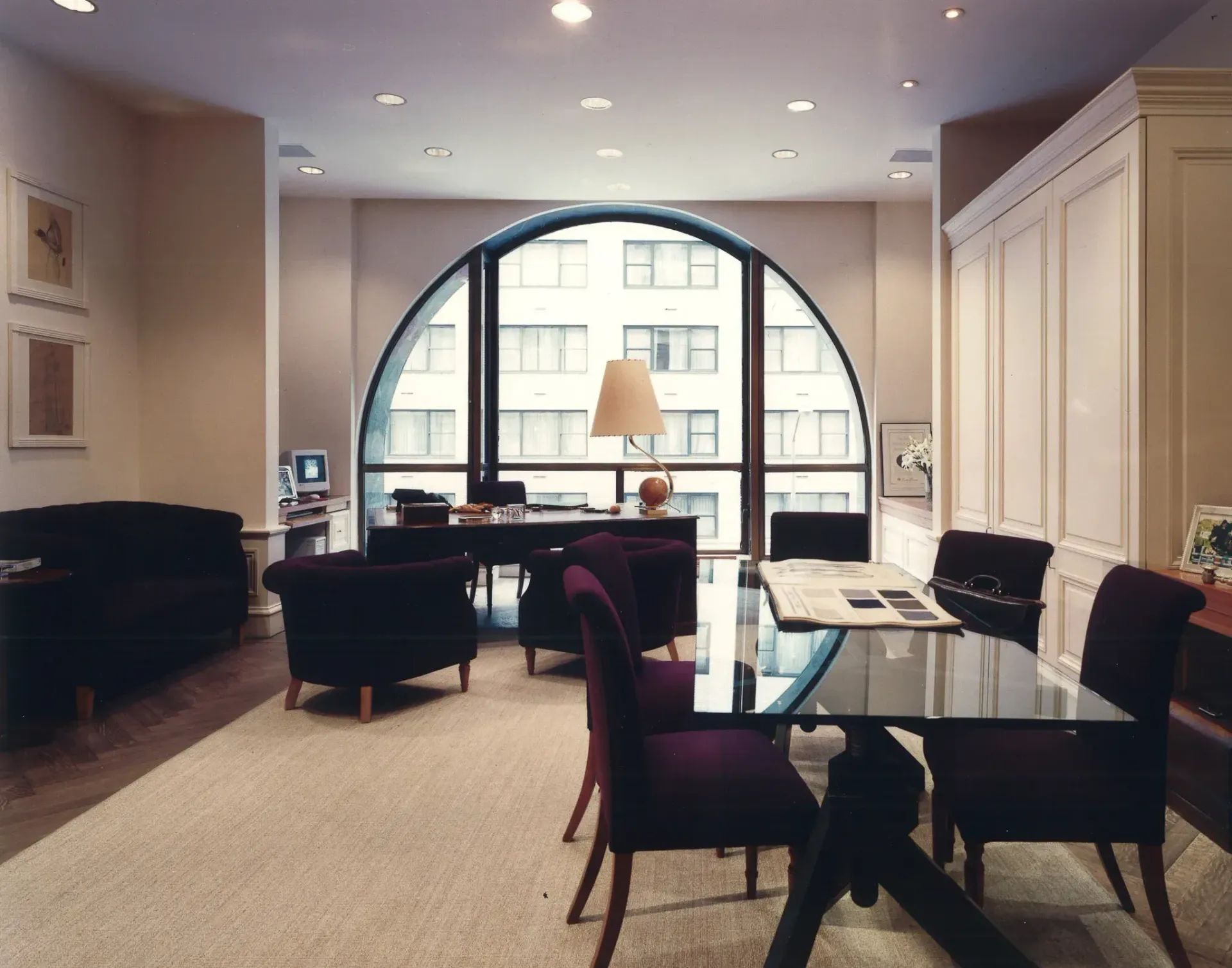Living room with arched window overlooking city buildings. Dark purple furniture and a glass table are visible.