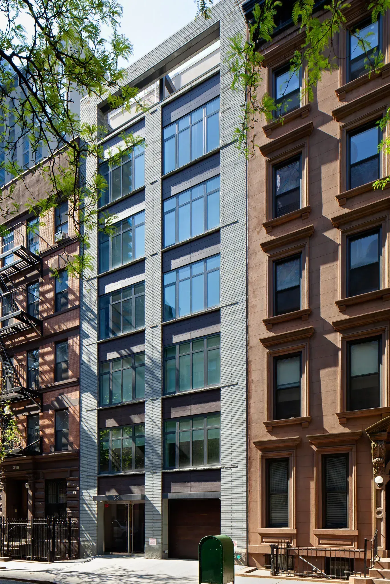 A modern, tall building with a gray facade stands between two brownstone buildings on a city street