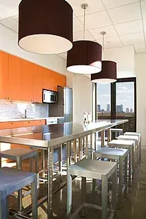 Modern kitchen with orange cabinets, stainless steel table and stools, and three pendant lights.