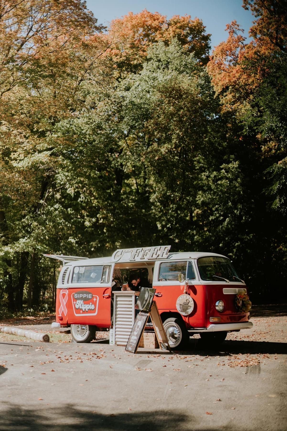 Red and white vintage VW bus with open side hatch. Man stands beside it, set in a wooded area with fall foliage.