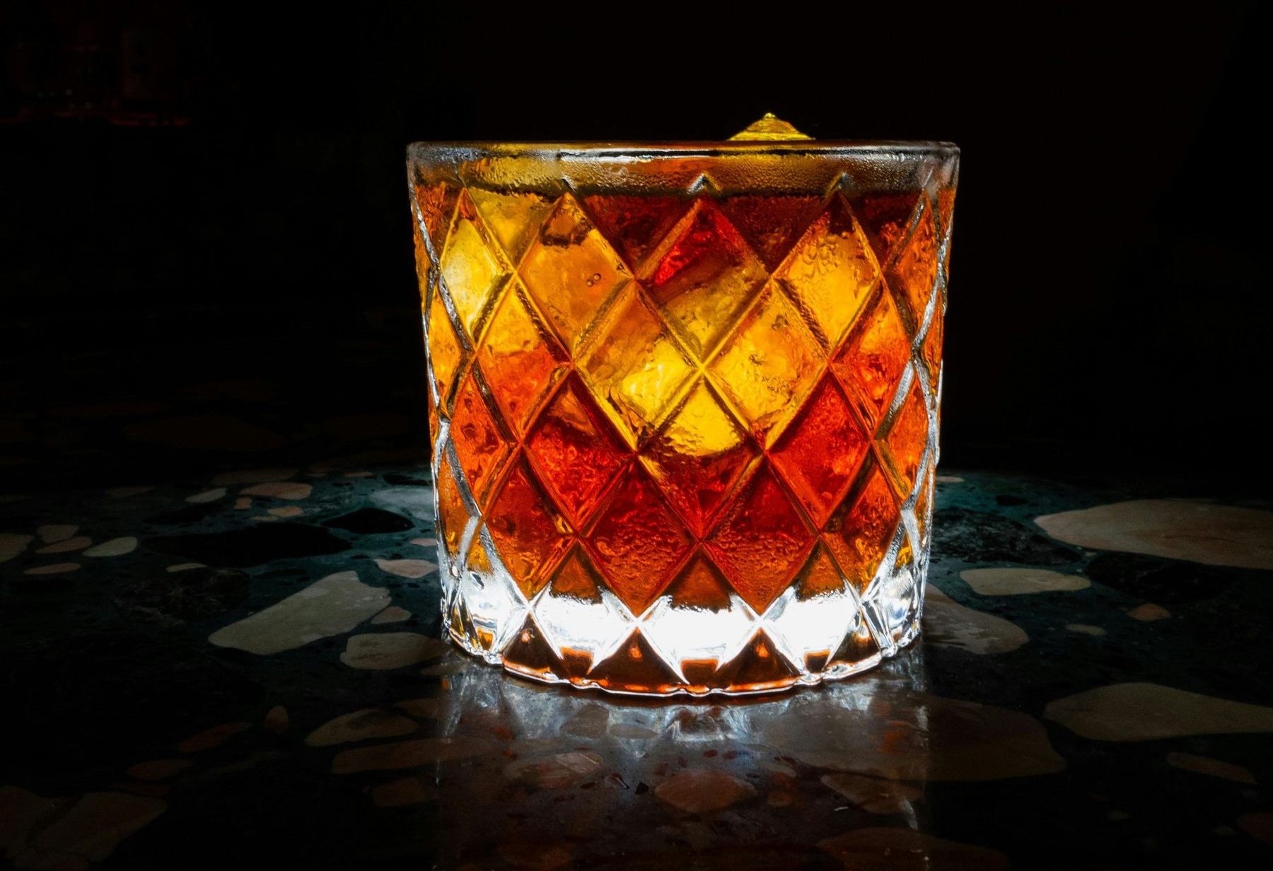 Cocktail in a diamond-cut glass, amber liquid with ice, on a dark countertop.